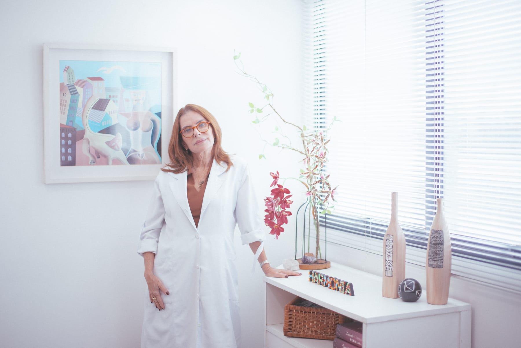 Dra. Katia Davy Bello de jaleco, em pé junto à janela do consultório, ao lado de um aparador com flores e um quadro na parede