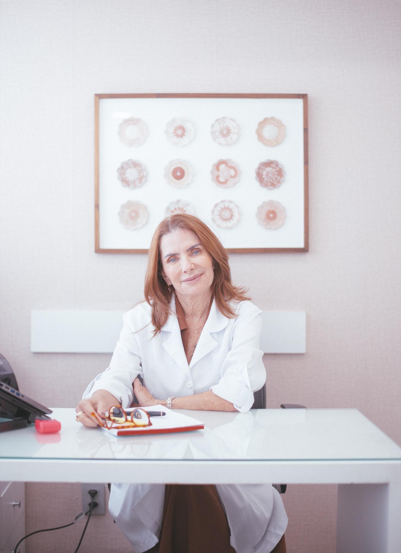 Dra. Katia Davy Bello no consultório, segurando os óculos sobre um caderno, com um quadro de flores emolduradas ao fundo