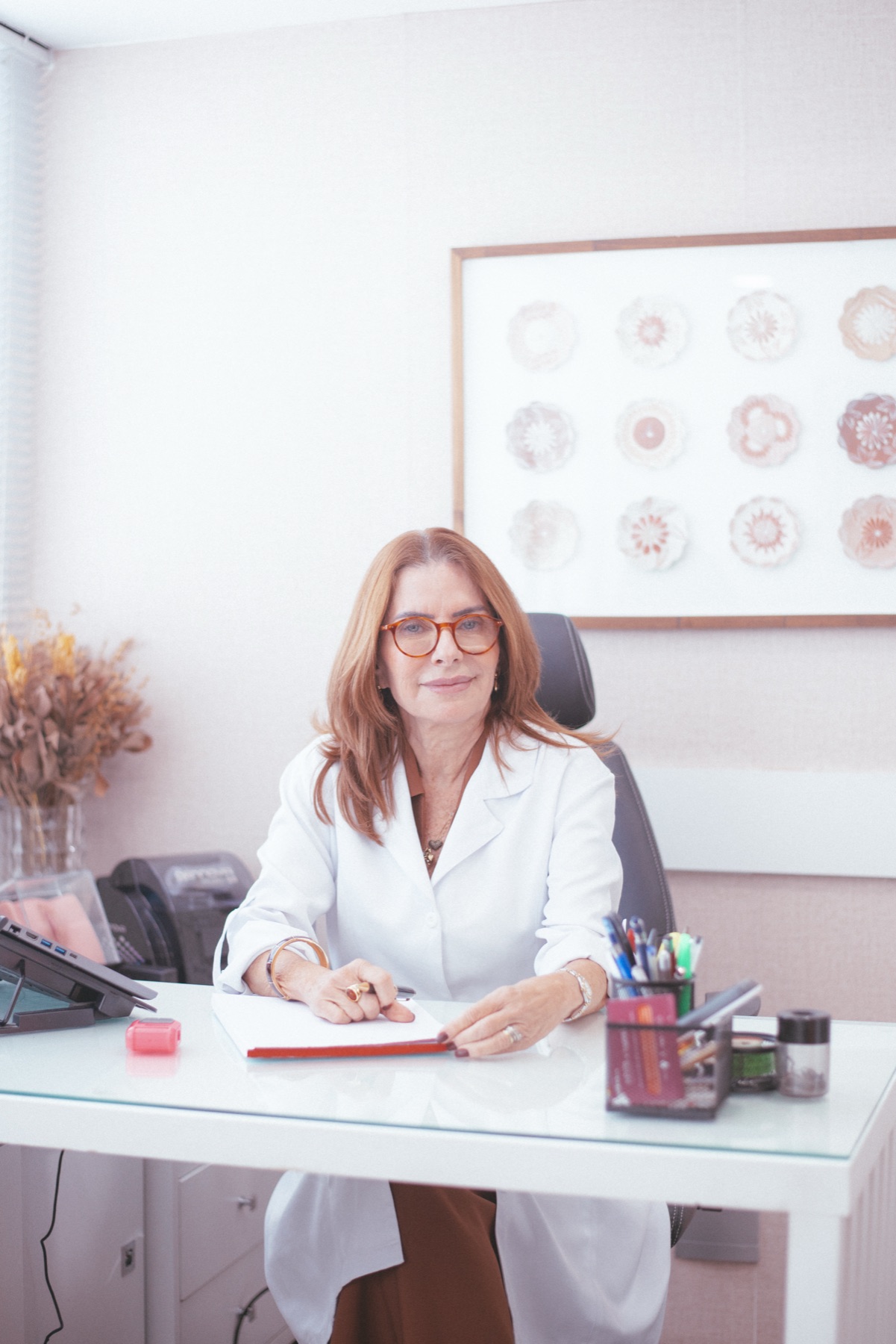 Dra. Katia Davy Bello sentada à mesa do consultório, de jaleco branco, escrevendo num caderno e olhando para a câmera